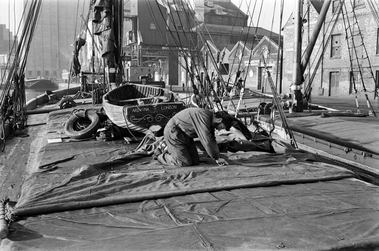 Black and white photograph showing a man leaning over on the deck of a Thames barge. Tarpaulin sheets cover the deck, and dock buildings can be seen in the background
