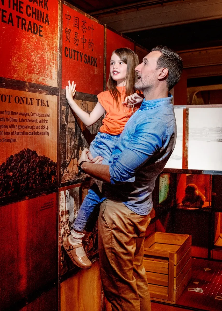 A father lifts his daughter up to look at a display of tea chests inside Cutty Sark