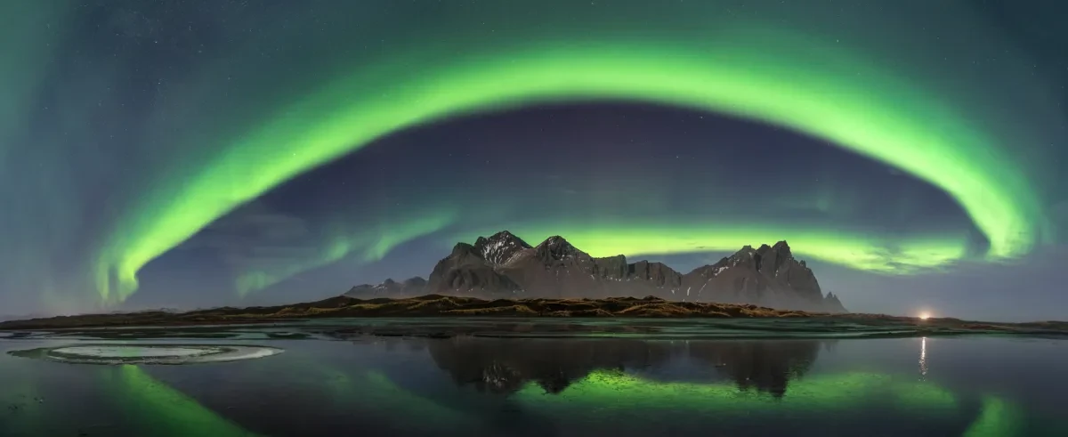 An arch of green light fills the night sky as the aurora appears over a calm lake. Mountains can be seen in the distance