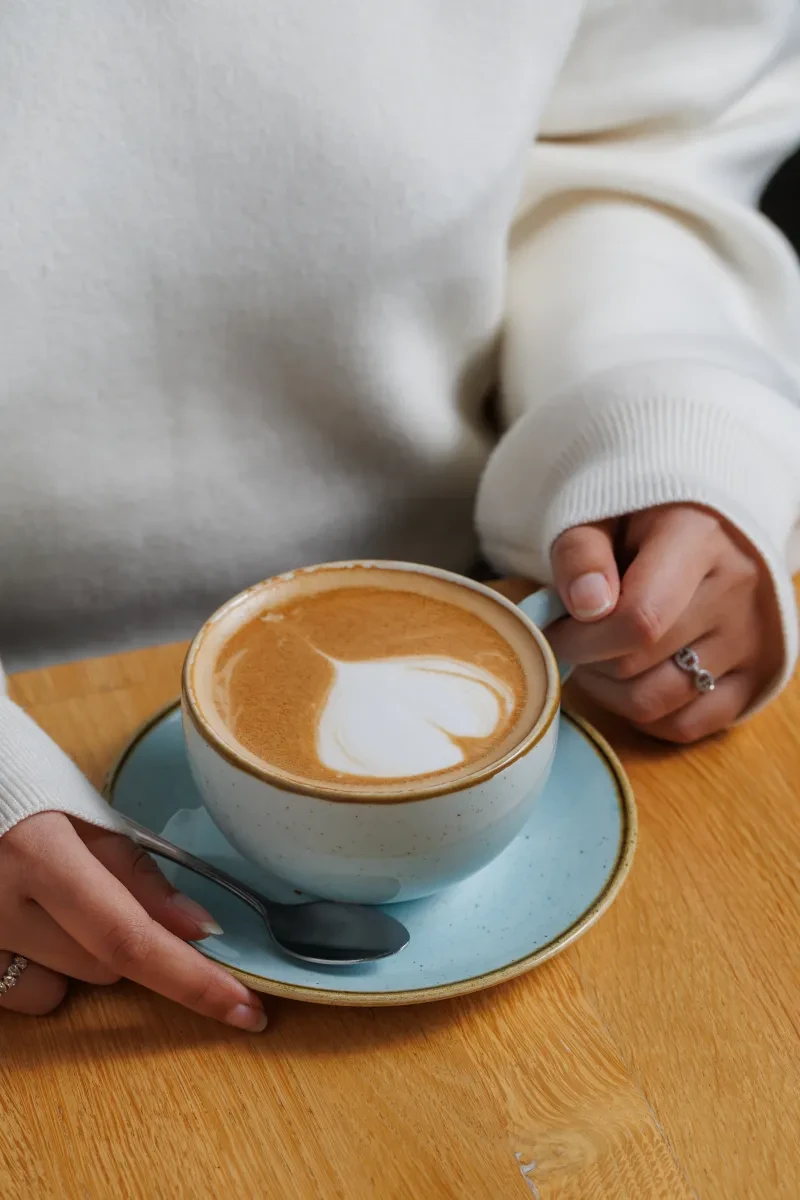 A coffee with a white foam heart on top, held by a woman wearing a snuggly white jumper