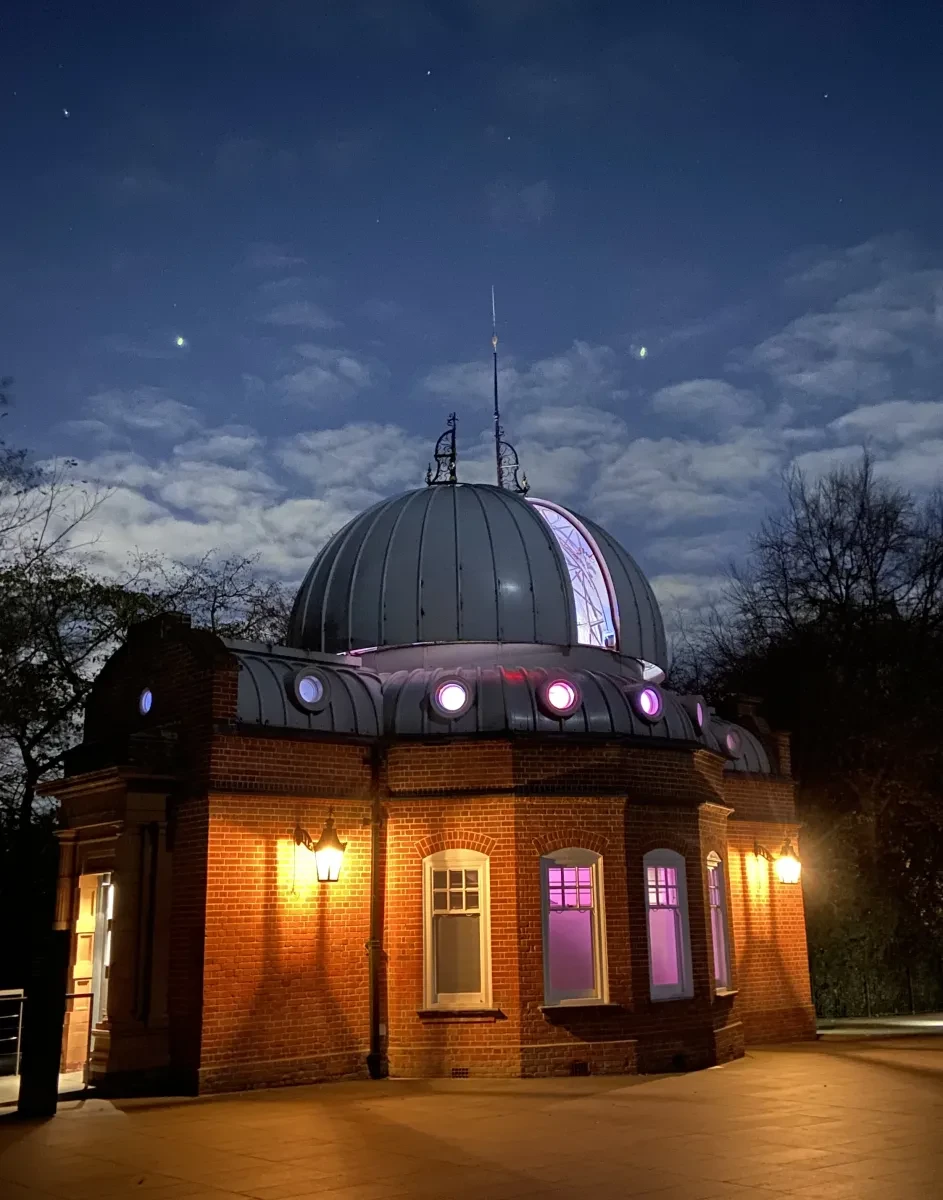 Photo of historic observatory building, the Altazimuth Pavilion, which is a small building with an observing dome on it. The dome is open and lit up in purple lights. There are stars in the sky overhead