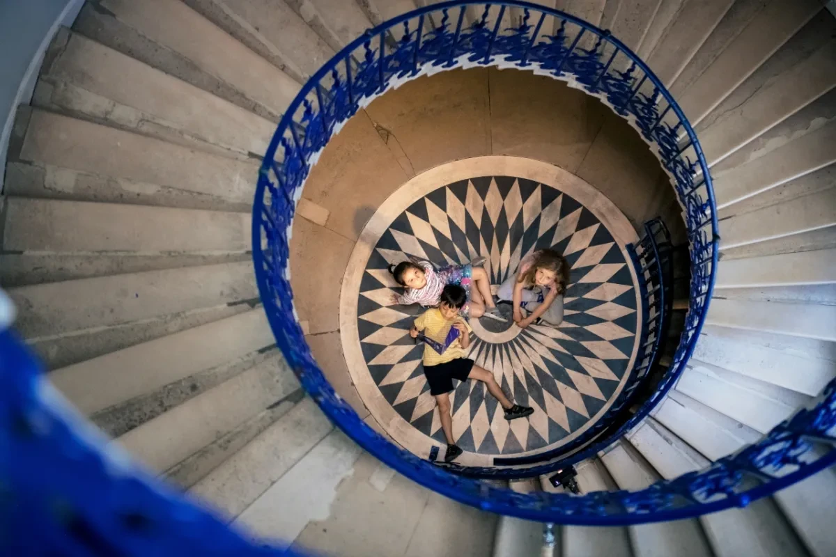 A group of children sit on the floor, looking up at a spiral staircase.