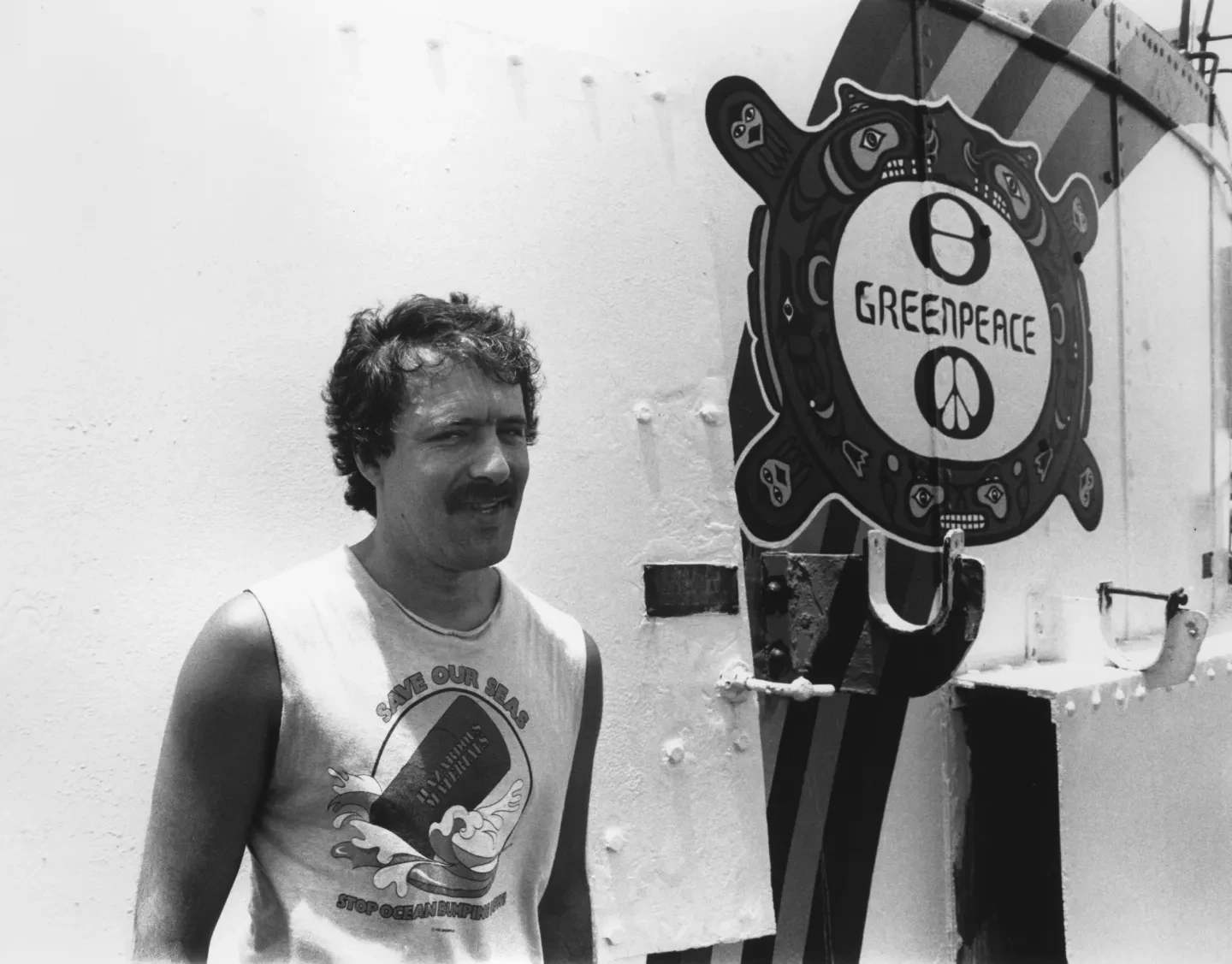 Black and white photograph of Greenpeace photographer Fernando Pereira, standing next to the ship Rainbow Warrior