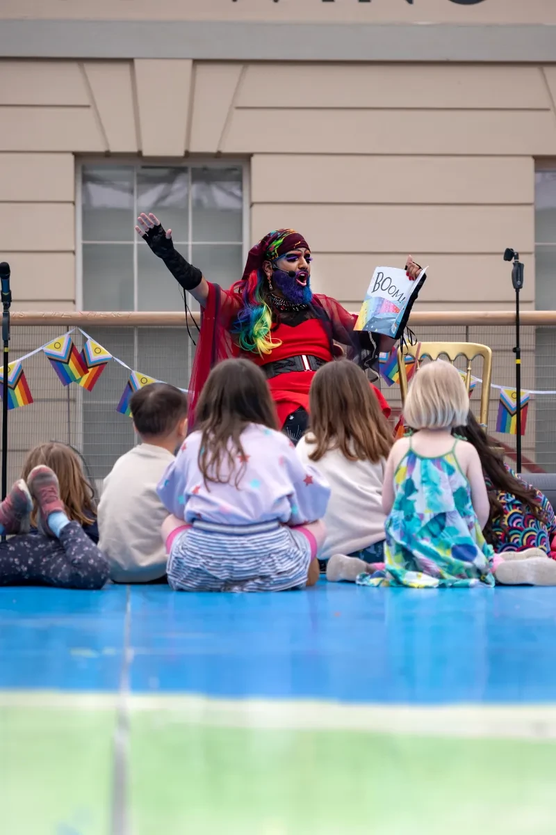 Photo of person in drag reading to children from a book in their hand and gesticulating