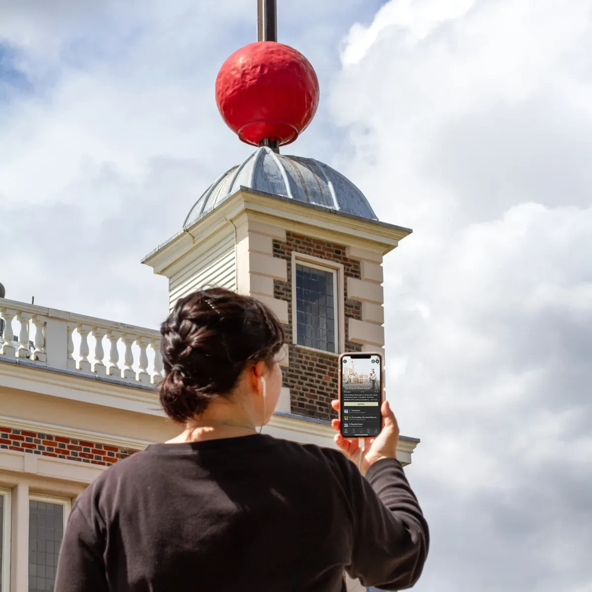 A woman holds her phone as she listens to the Royal Observatory audio tour, with the Observatory buildings in the background