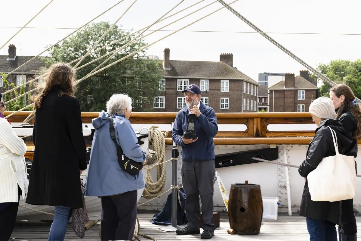 Photo of a man in a navy jacket holding up a historic item and smiling as a group of people listening to him. They are on the deck of a historic ship