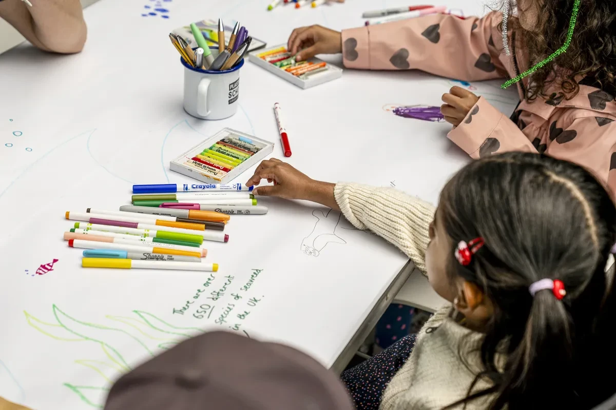 Children sitting at a table covered by a large white sheet of paper. Pens and pencils are strewn across it, ready for the kids to take part in a craft activity
