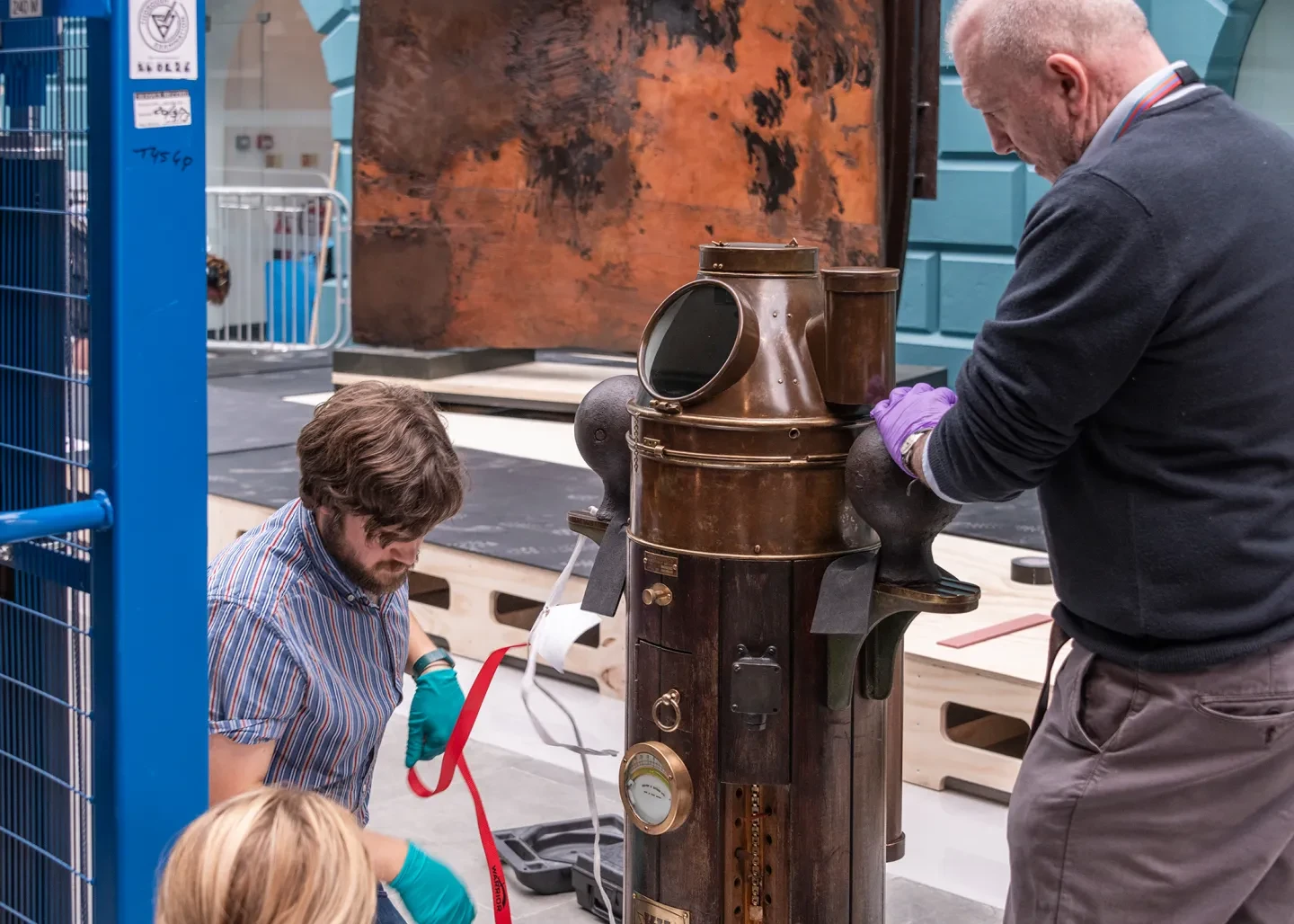 Museum object handlers work on the ship's binnacle from Rainbow Warrior at the National Maritime Museum. The metal object is large and cone-shaped, with a hole where the compass would have been mounted